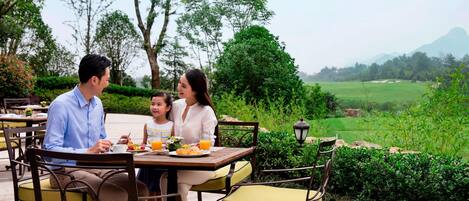 Una familia cena al aire libre en una mesa con sillas, rodeada de follaje y una vista distante de colinas.