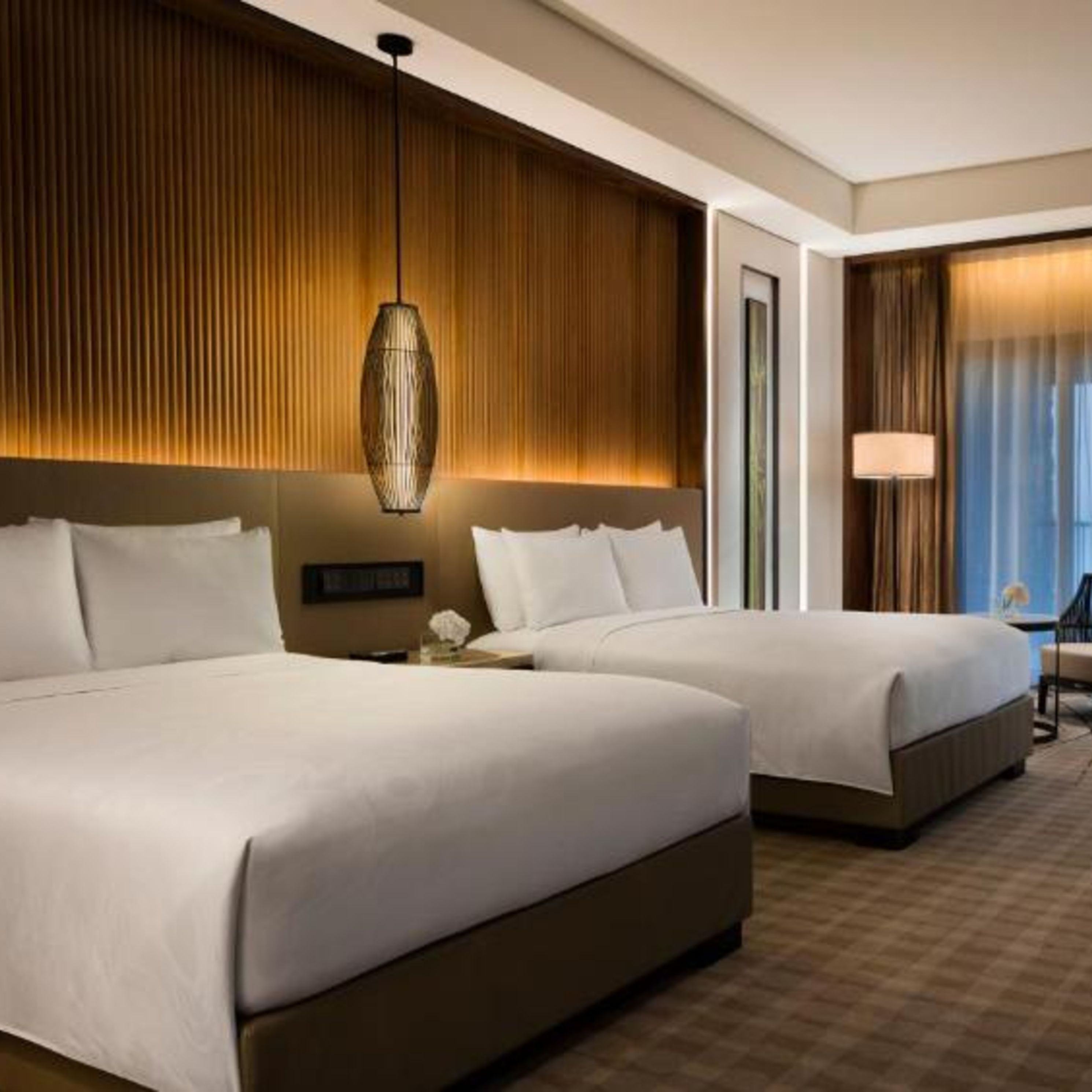 A hotel room with two beds, a wooden panel wall, a hanging light fixture, and a view of the outside through a window with curtains.