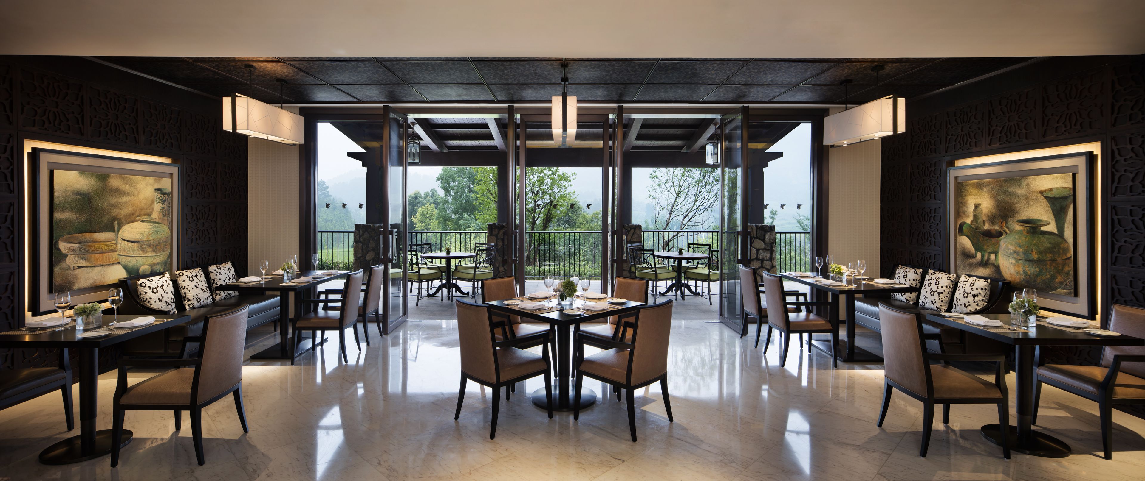 A spacious dining area with multiple tables and chairs, large windows offering a view of trees and mountains, and decorative wall art.