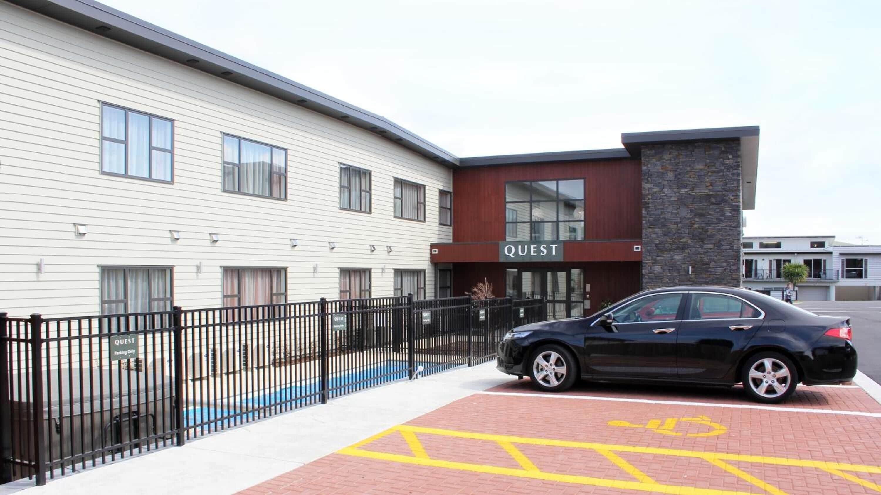 A modern building with a 'QUEST' sign, a black car parked in front, and a wheelchair-accessible parking space.