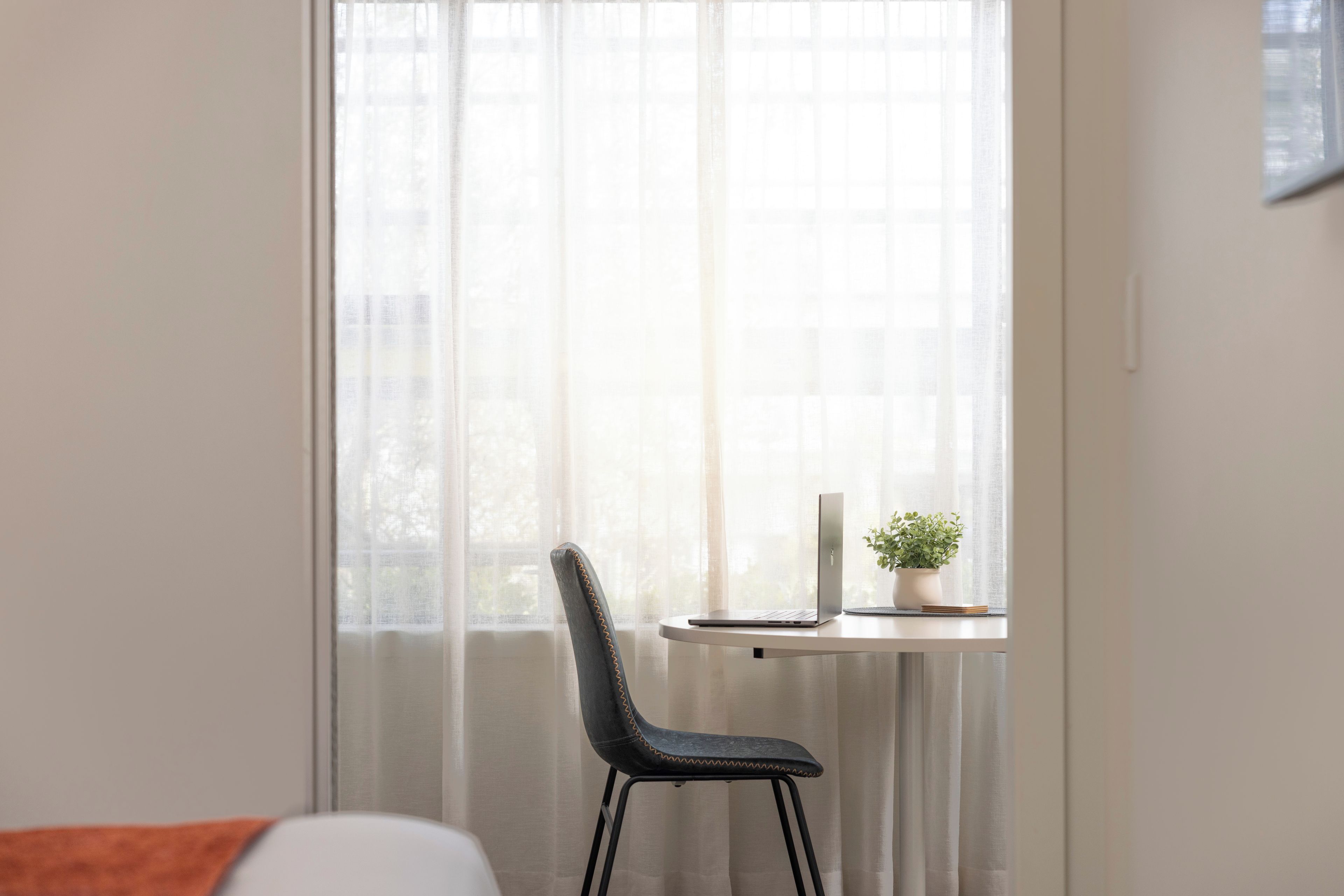A room with a single window, a chair, a small table with a laptop and a plant.