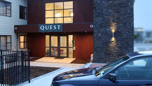 Un bâtiment moderne avec une enseigne « QUEST », une façade en pierre et une voiture garée devant.