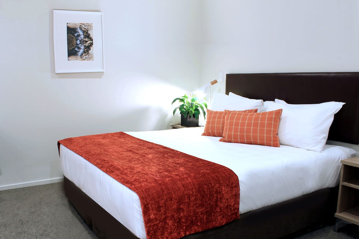 Een net opgemaakt bed met wit-oranje beddengoed, een bruin hoofdbord en een rode plaid.