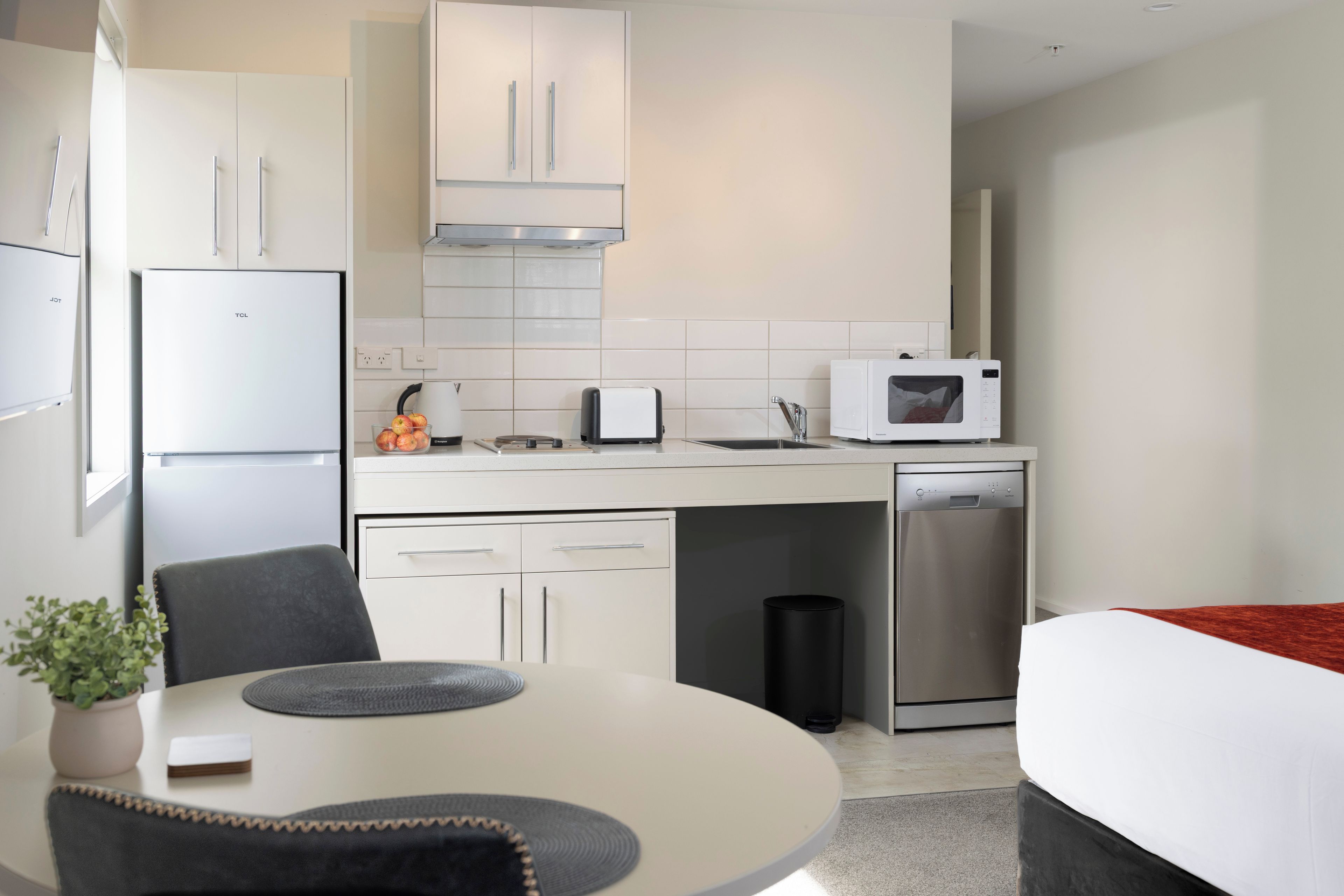 Una cocina moderna con armarios blancos, microondas, tostadora y lavavajillas. Hay una mesa de comedor con sillas y una pequeña planta en maceta.