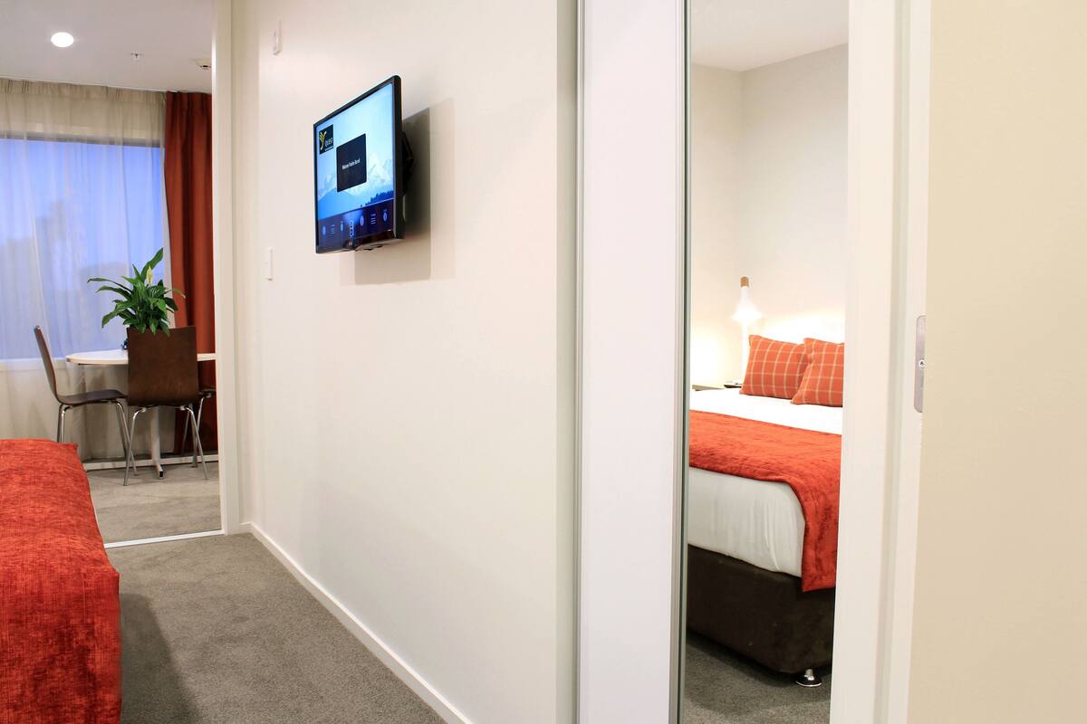 Een hotelkamer met een bed, een bureau met stoel, een aan de muur bevestigde televisie en een bloempot.