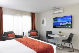 Quarto de hotel com cama, duas poltronas, uma mesinha e uma televisão exibindo uma mensagem de boas-vindas.