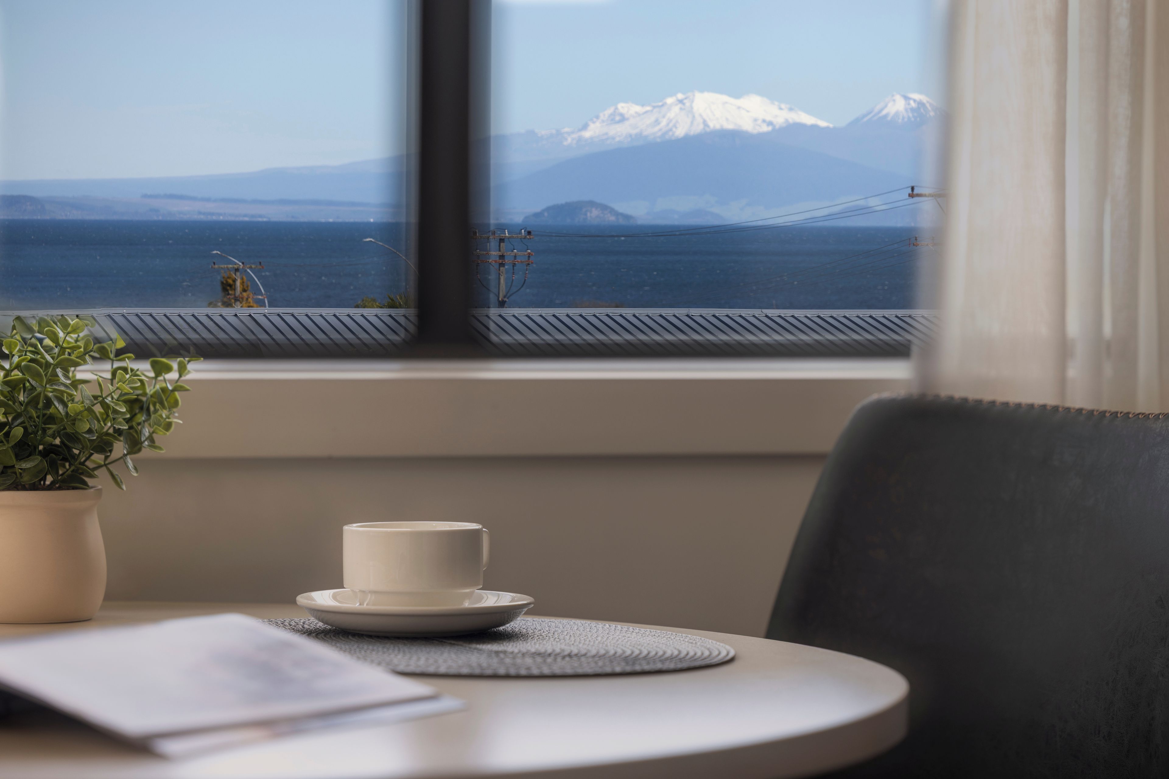 Ein Tisch mit Tasse und Untertasse, einer Topfpflanze und Blick auf eine Berglandschaft und eine Brücke durch ein Fenster.
