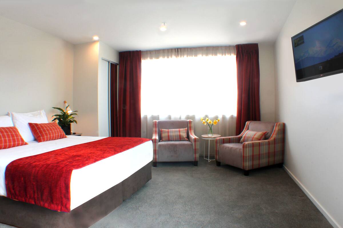 Een hotelkamer met een groot bed, twee fauteuils, een tafeltje met bloemen en een televisie aan de muur.