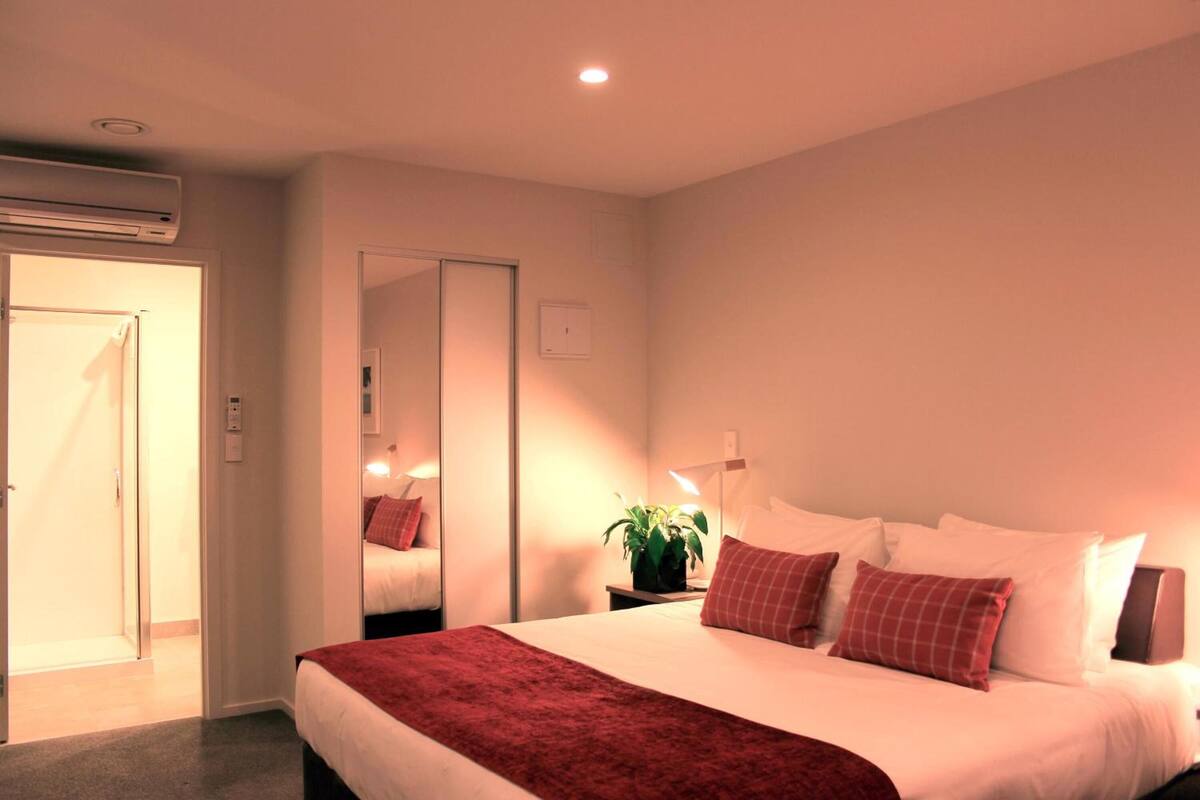 Een hotelkamer met een bed, nachtkastje met lamp, een potplant en een spiegel.