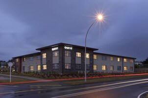 Um edifício moderno com a sinalização "QUEST", várias janelas e uma rua com rastros de luz.