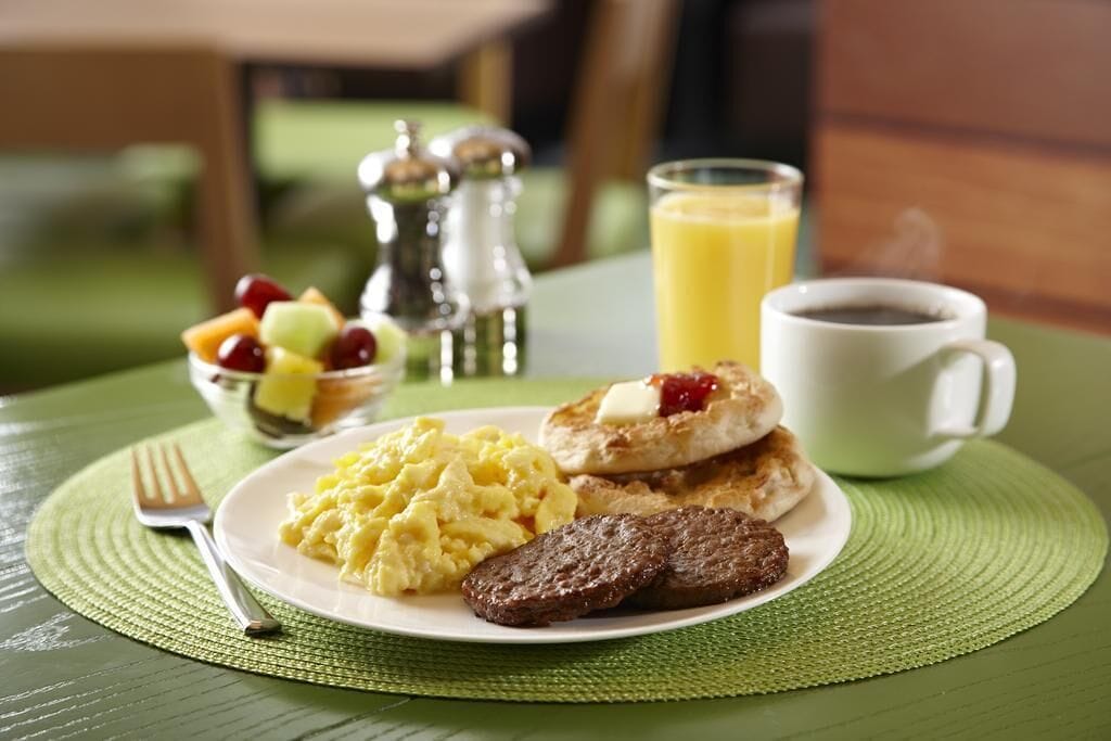 Une assiette de petit-déjeuner comprenant des œufs brouillés, des saucisses et des toasts à la confiture, accompagnée d’un verre de jus d’orange et d’une tasse de café.