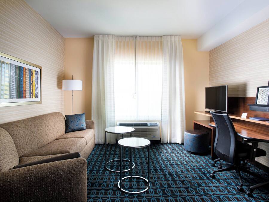 Quarto de hotel com sofá, mesinha redonda, escrivaninha com cadeira, televisão e uma janela grande com cortinas.