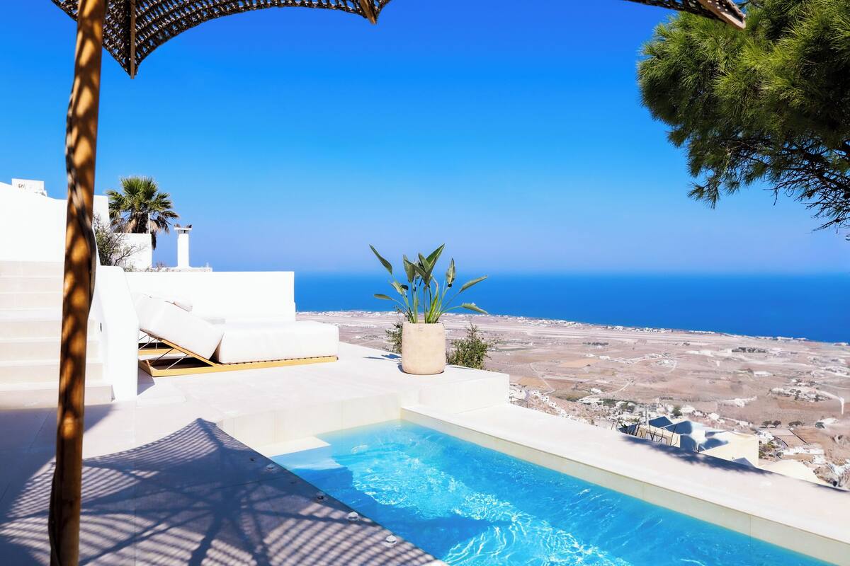 Ein Poolbereich mit freiem Blick auf das Meer, weißen Gartenmöbeln und einer Kühlpflanze.