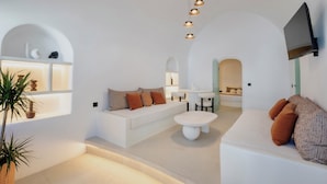 A modern living room with a white sofa, a coffee table, and a TV mounted on the wall. - Luna Santorini Suites (Santorini)