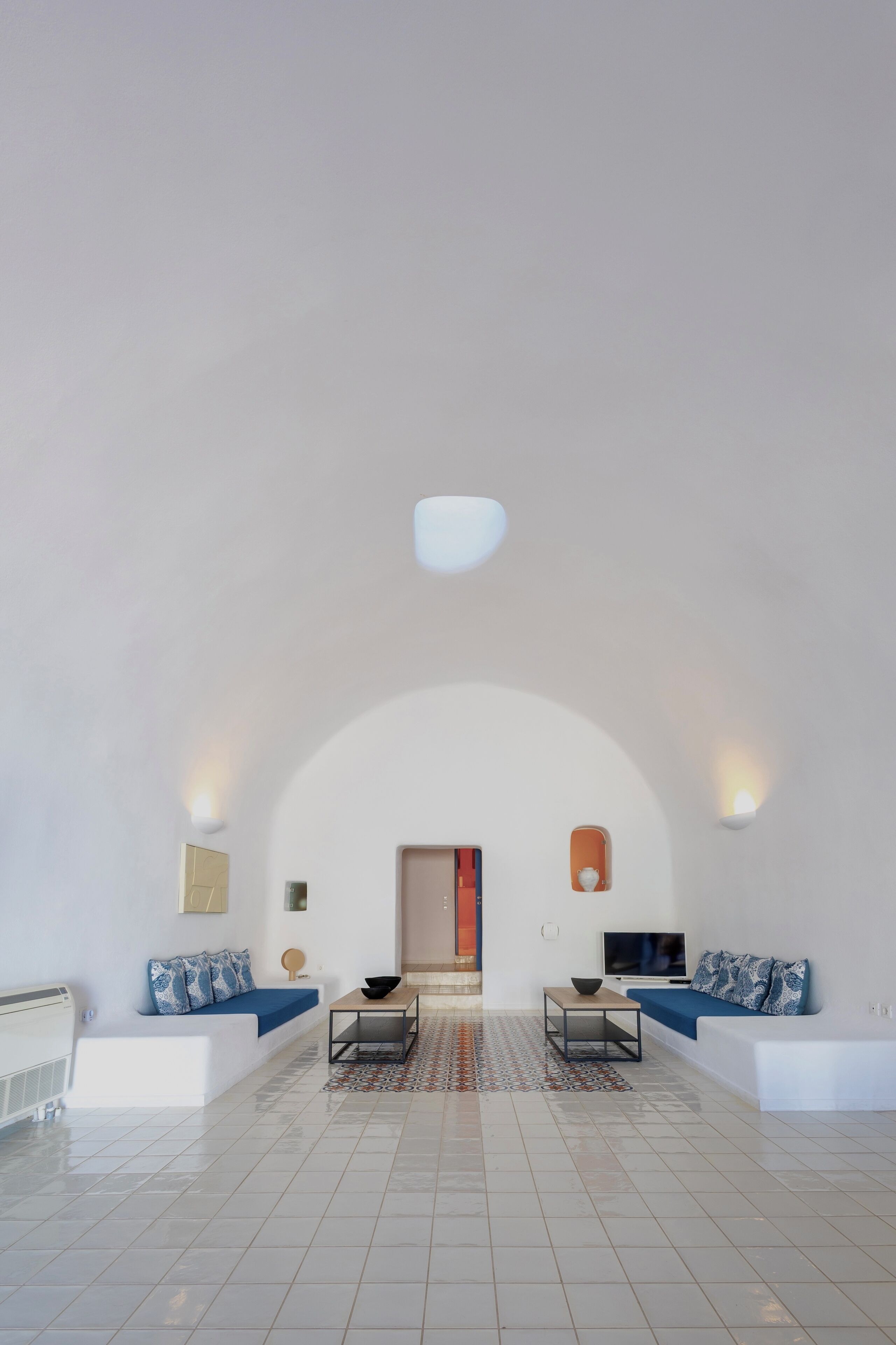 A spacious living area with a high ceiling, white walls, and a tiled floor. There are two sofas with blue cushions, a coffee table, and a television.