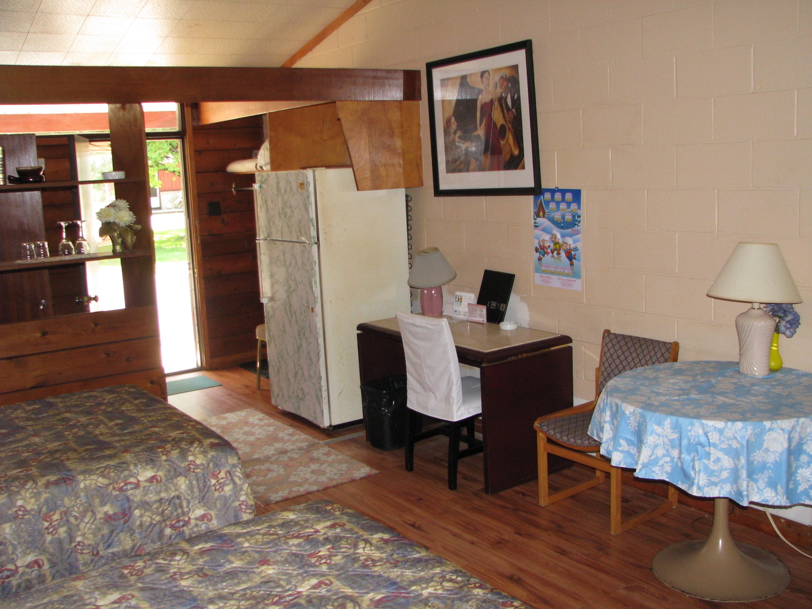 Una habitación pequeña y acogedora con dos camas, un escritorio, una silla, una mesa, un refrigerador y una lámpara.
