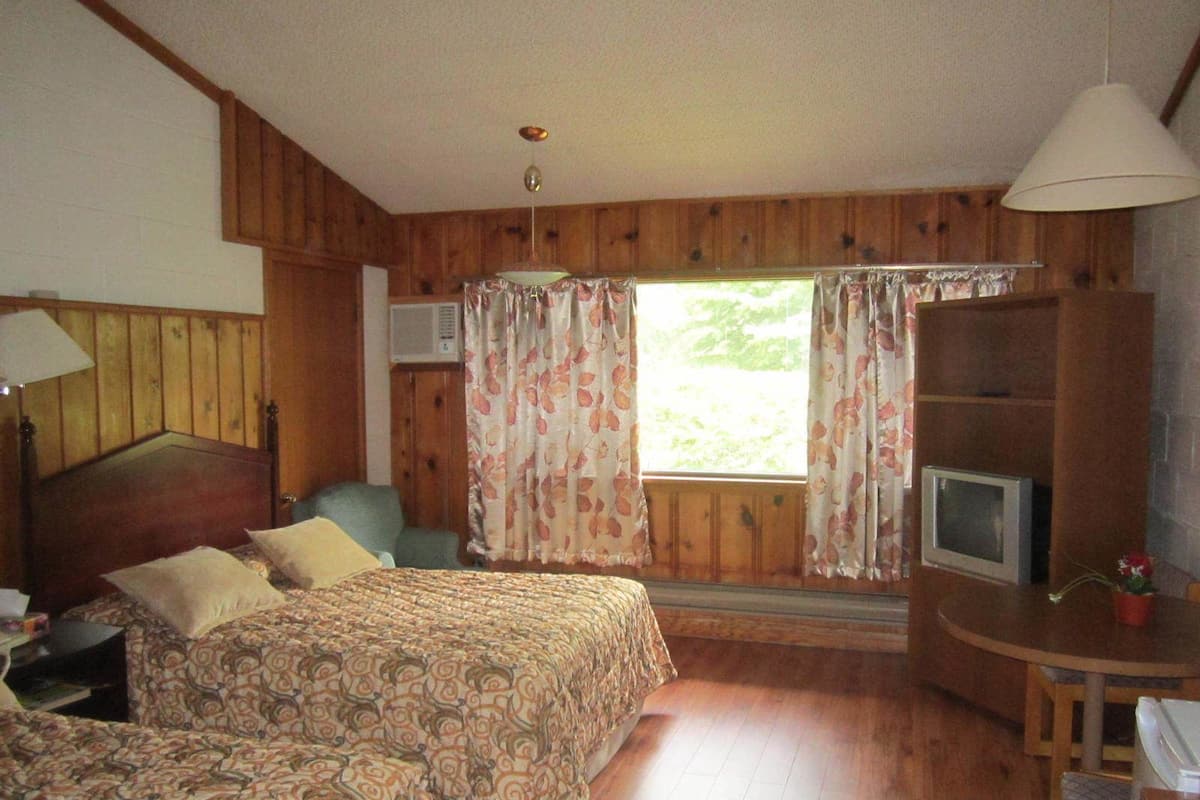 Ein Zimmer mit Holzwandverkleidung, einem Bett mit gemusterten Betttextilien, einem Schreibtisch mit Fernseher und einem fenster mit Vorhängen.