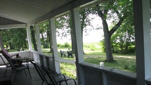 A covered patio with a table and chairs, overlooking a grassy area with trees and a bench.