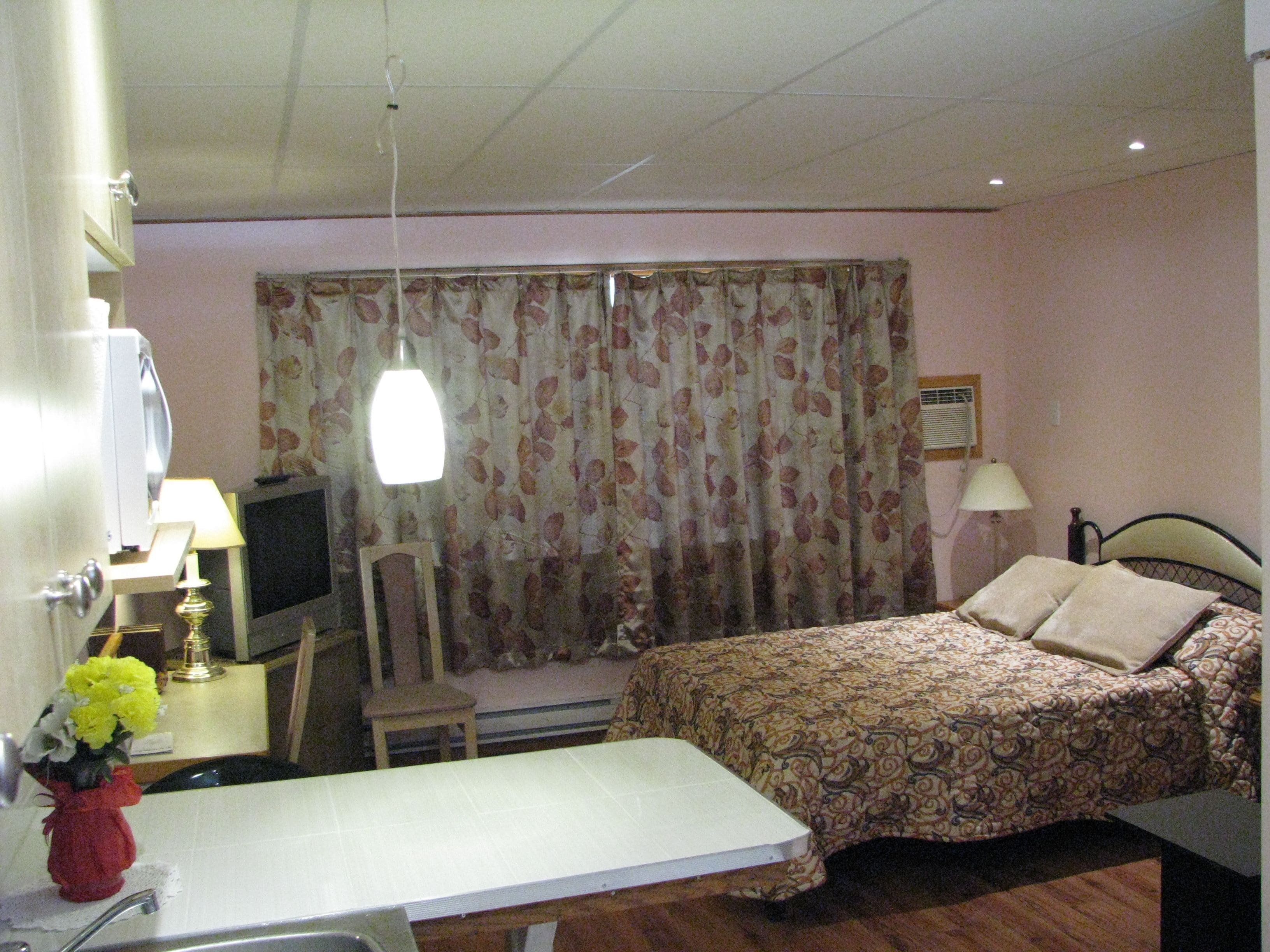 Una habitación de hotel con cama, escritorio con lámpara, una silla, televisor y ventana con cortinas.