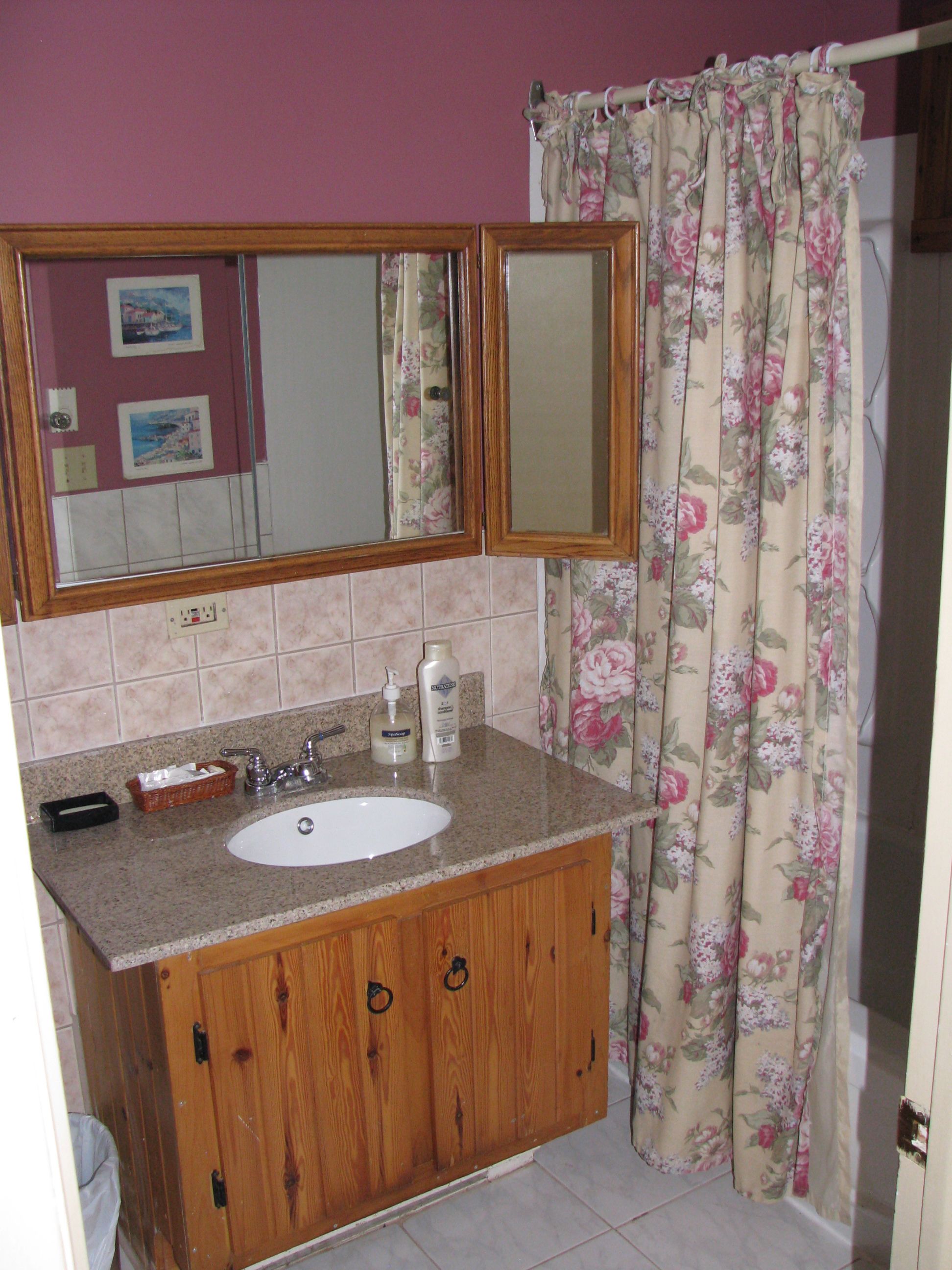 Un baño con tocador de madera, lavamanos blanco, espejo sobre el lavamanos y cortina de ducha con estampado floral.