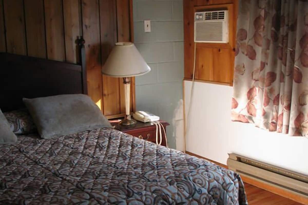 Ein Schlafzimmer mit einem geblümten Bettbezug, einer Nachttischlampe, einem Telefon und einem Fenster mit floralen Vorhängen.