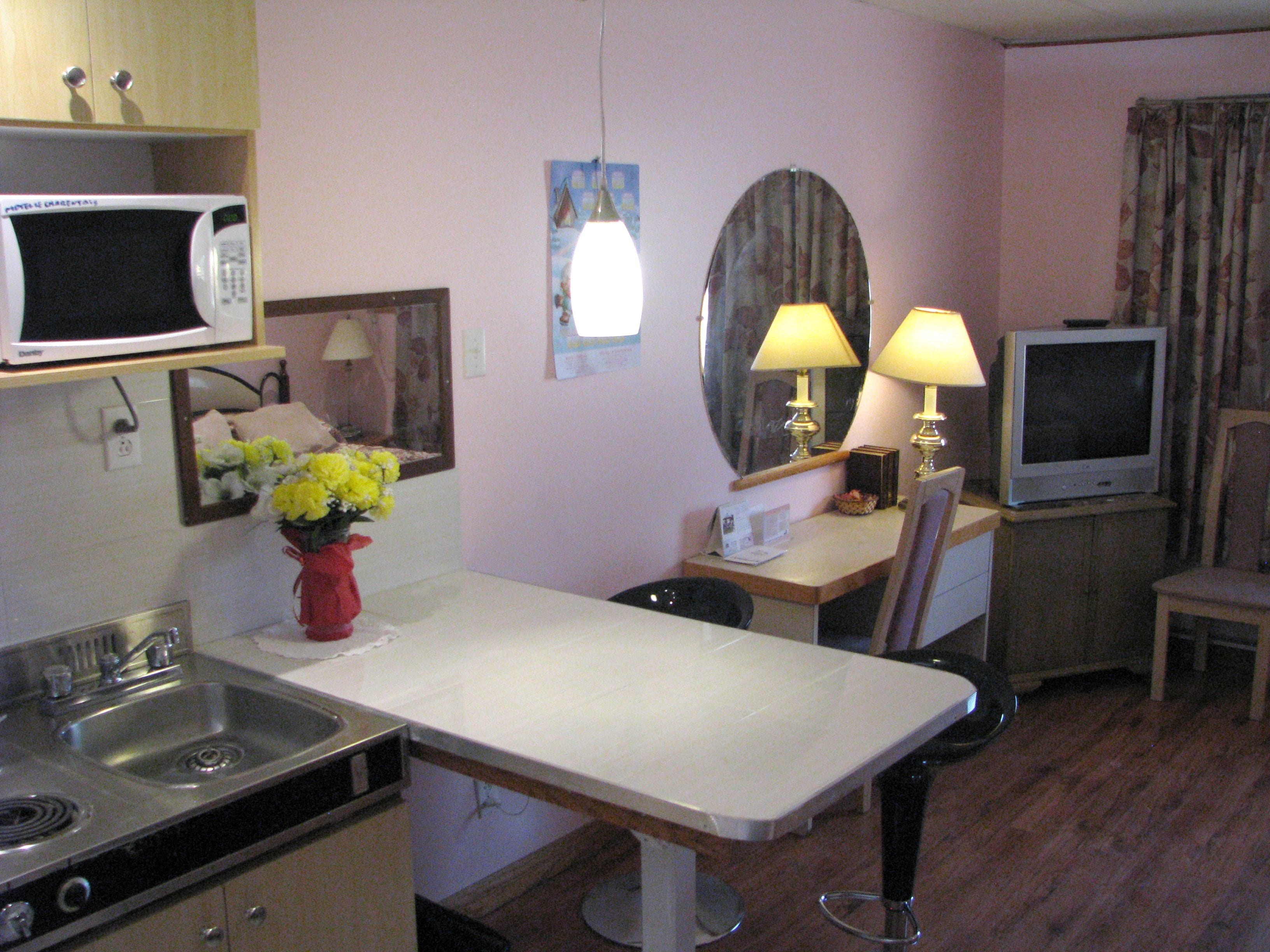 A compact kitchen with a microwave, sink, and stove. There is a small table with a chair, a TV, and a lamp.