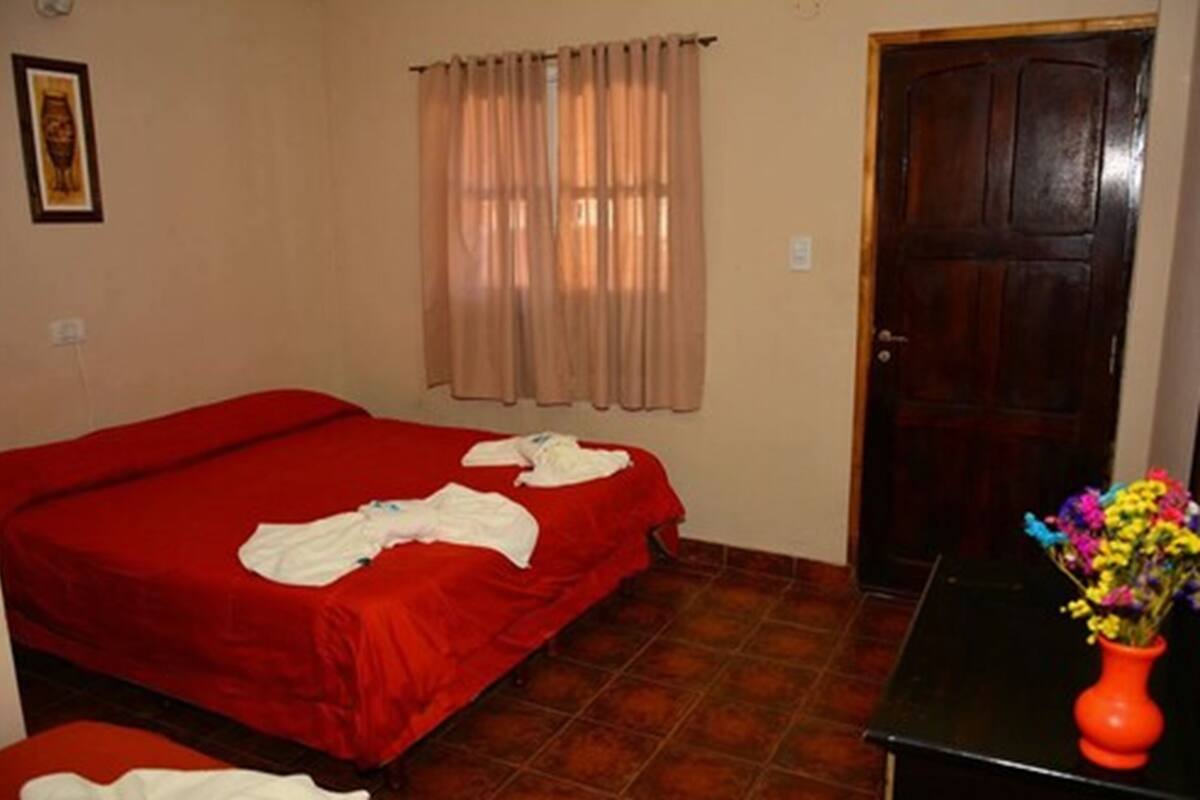 Una habitació amb dos llits, una porta de fusta, una finestra amb cortines i un vas amb flors sobre una taula.
