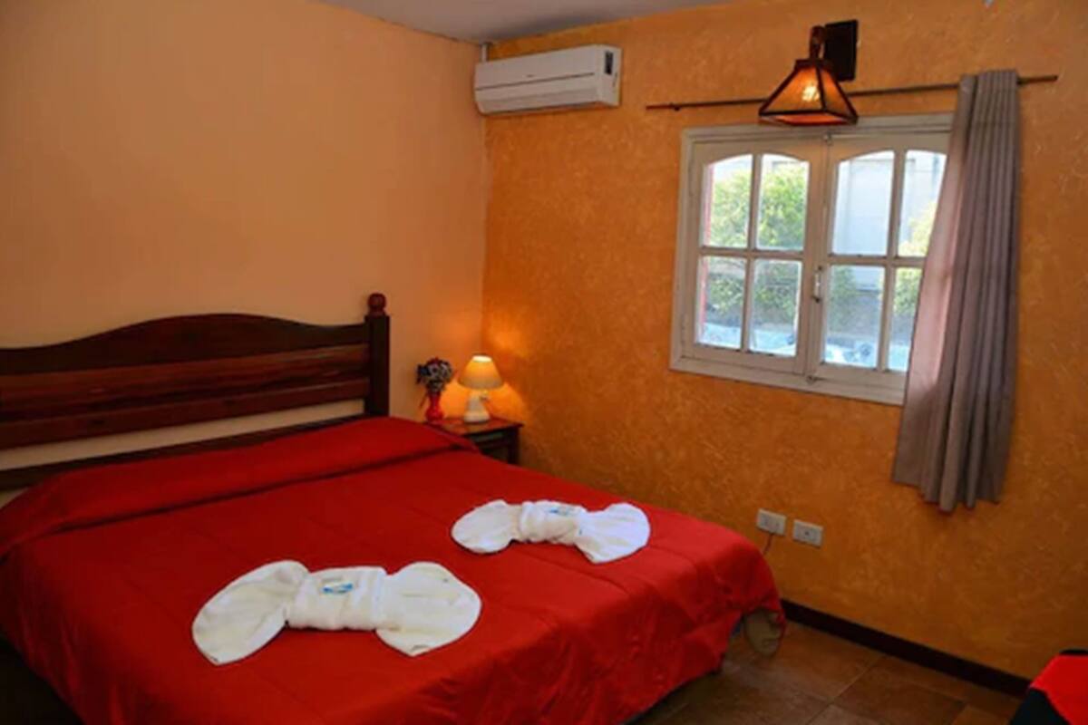 Habitació d'hotel amb llit vermell, finestra amb cortines, aire condicionat i làmpada sobre una taula de nit.