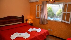 Habitación de hotel con colcha roja, cabecero de madera, ventana con cortinas y lámpara en la mesita de noche.