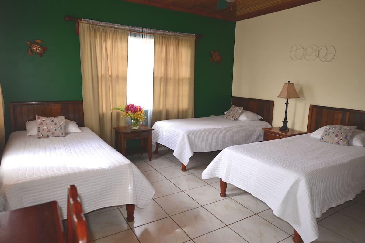A hotel room with three single beds, a wooden headboard, a small table with a vase of flowers, and a lamp.