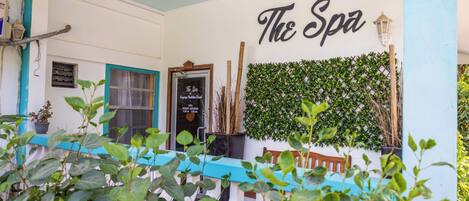 Un spa con un letrero que dice “The Spa” sobre un muro de follaje, un banco de madera y una mesa con mantel azul.