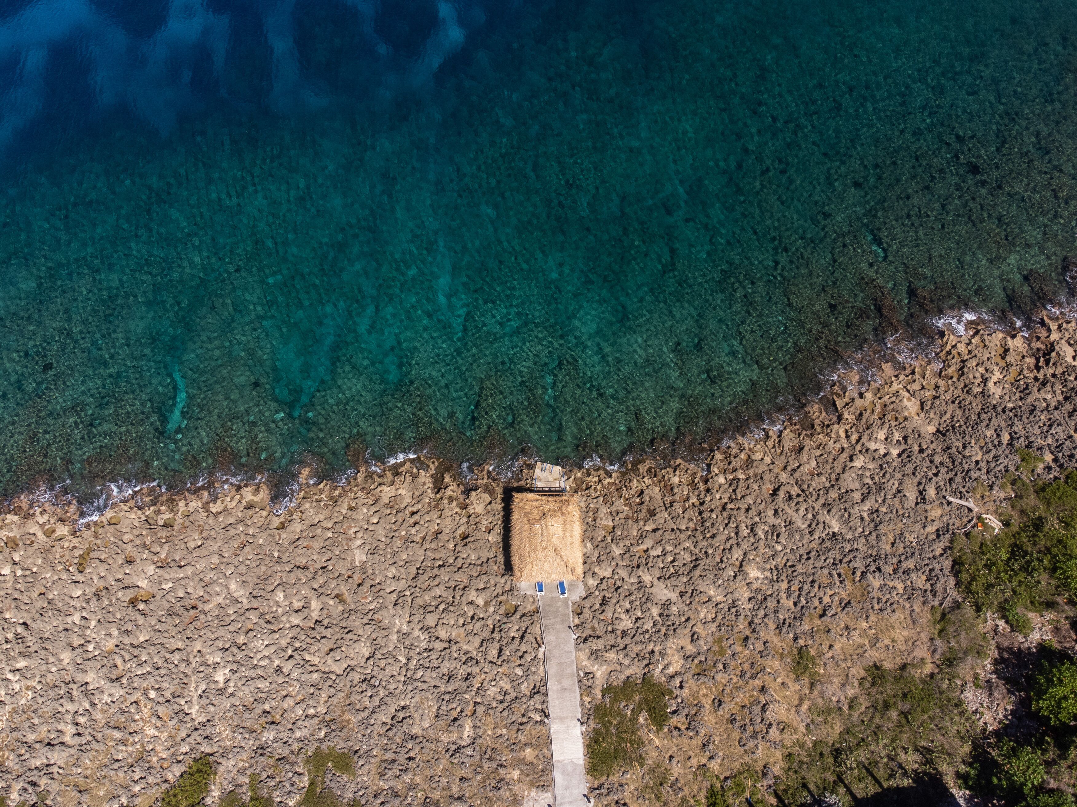 An aerial view of a coastal area with a rocky shoreline, a narrow concrete pathway leading to a building, and clear blue water.
