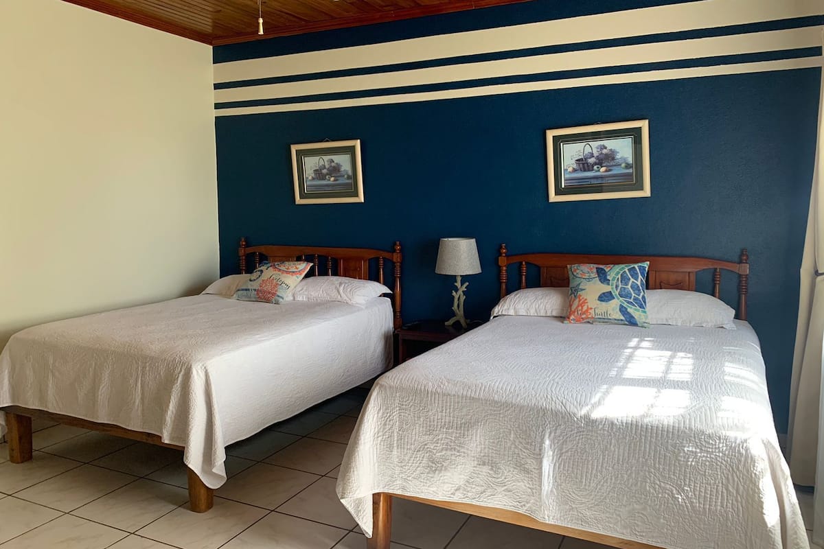 A hotel room with two single beds, wooden headboards, white bedding, and a ceiling fan.