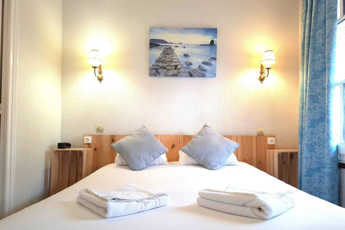 Une chambre d’hôtel avec un lit, deux lampes fixées au mur, un tableau et une fenêtre avec des rideaux.