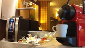 A coffee machine and a coffee maker on a countertop with a cup of coffee, a tray of pastries, and a small statue. - Hôtel Le Chantilly (Deauville)