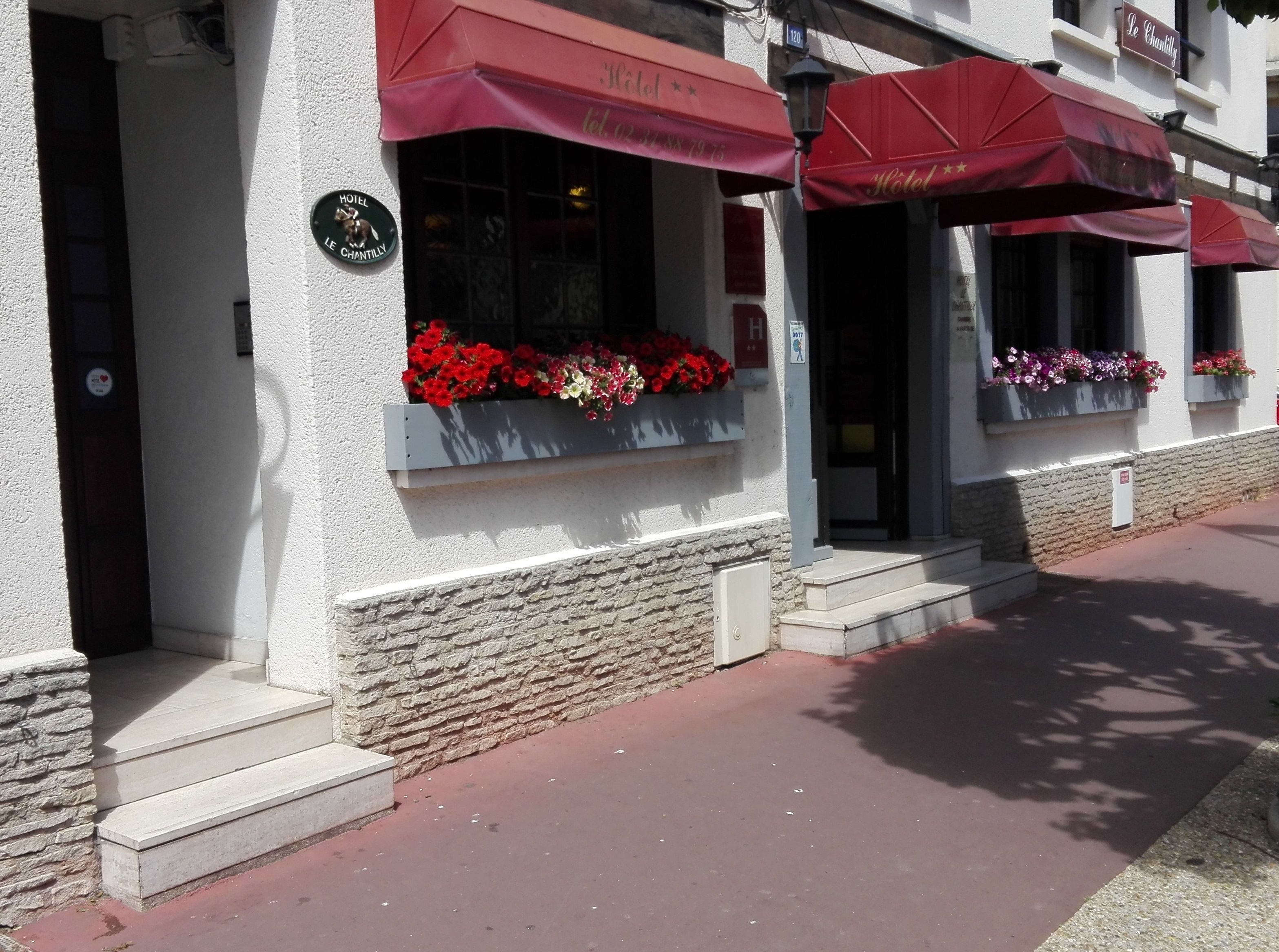 A hotel facade with red awnings, flower boxes with red and pink flowers, and a stone wall.