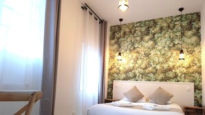 A bedroom with a bed, two bedside tables, a chair, a window with curtains, and a wall with a floral mural. - Hôtel Le Chantilly (Deauville)