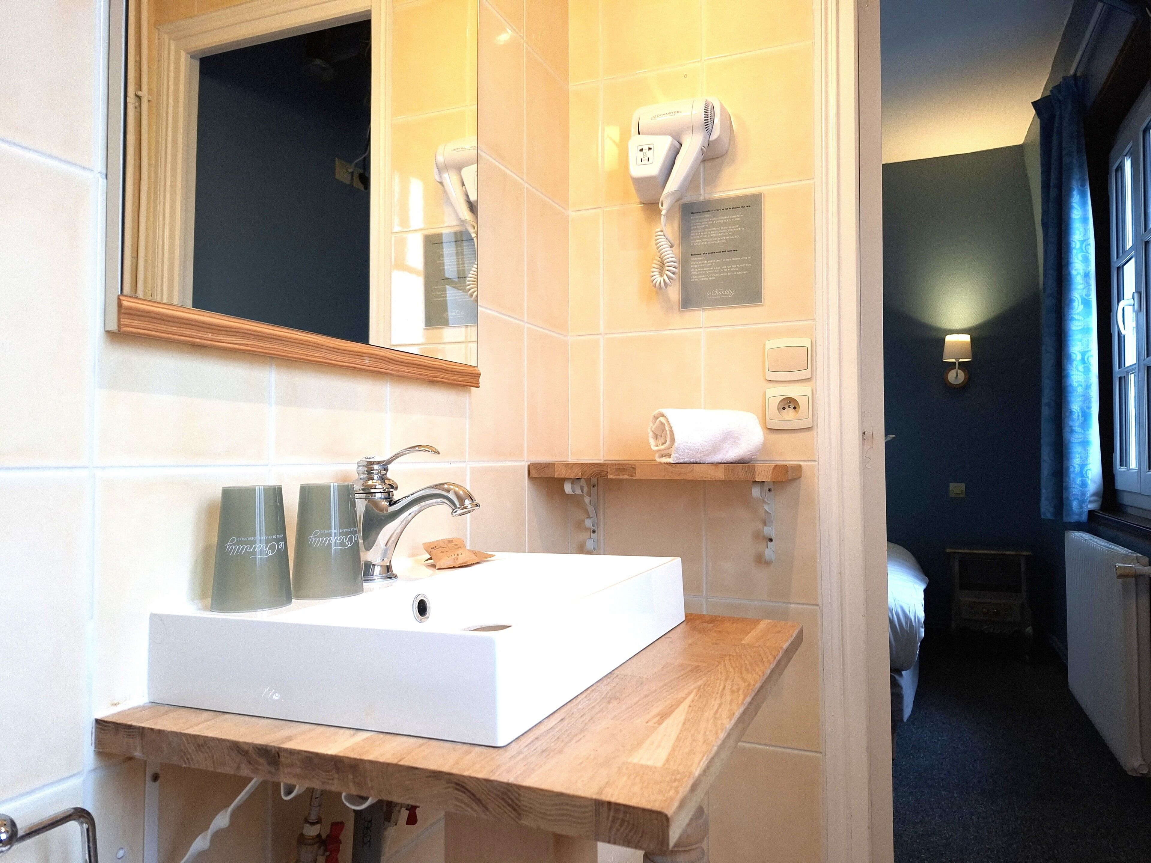 Une salle de bain avec un lavabo blanc, un miroir, un sèche-cheveux et une serviette posée sur une étagère.