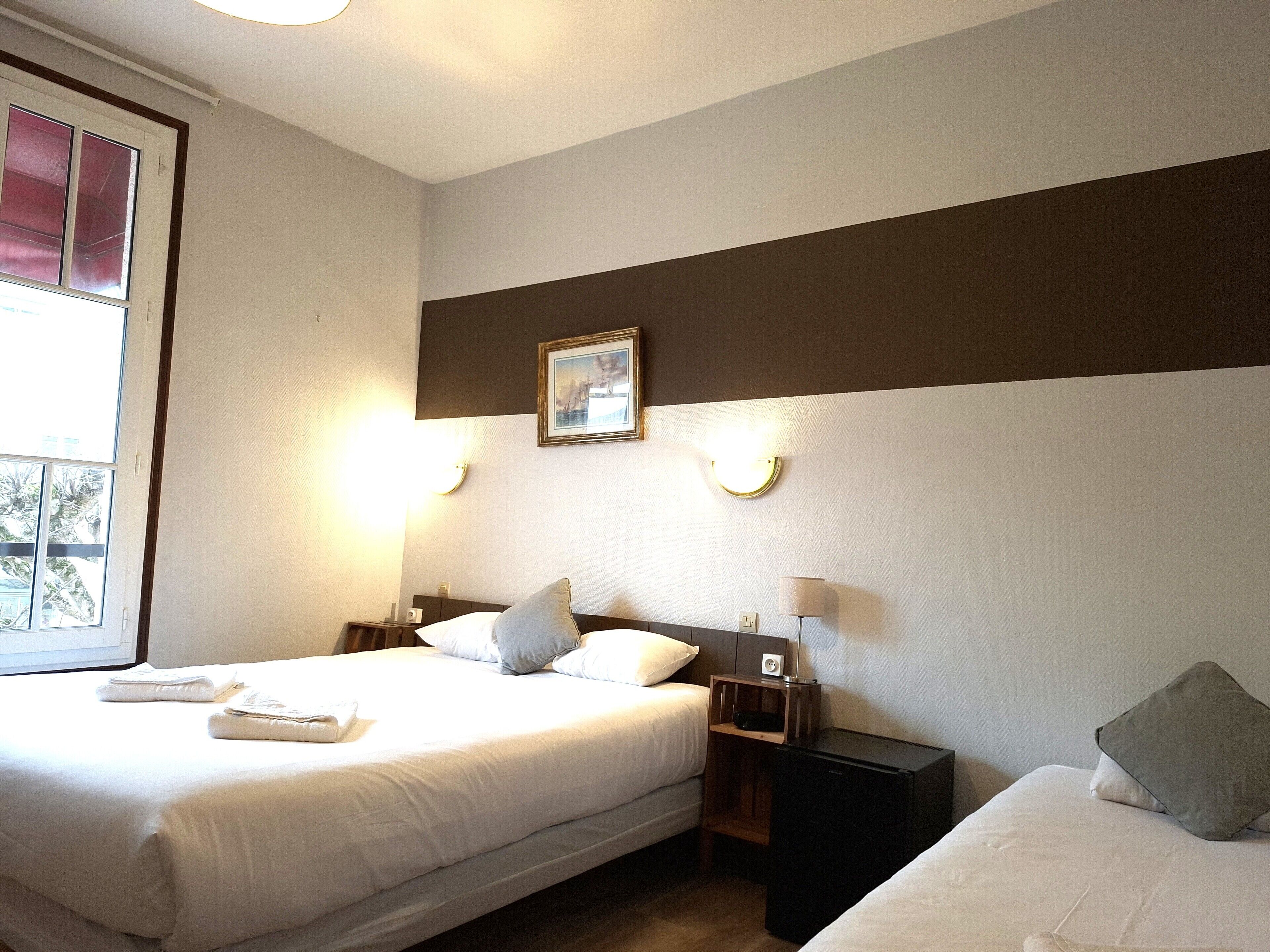 Habitación de hotel con dos camas, un escritorio pequeño, una mesita de noche, un cuadro enmarcado y una ventana con persianas.