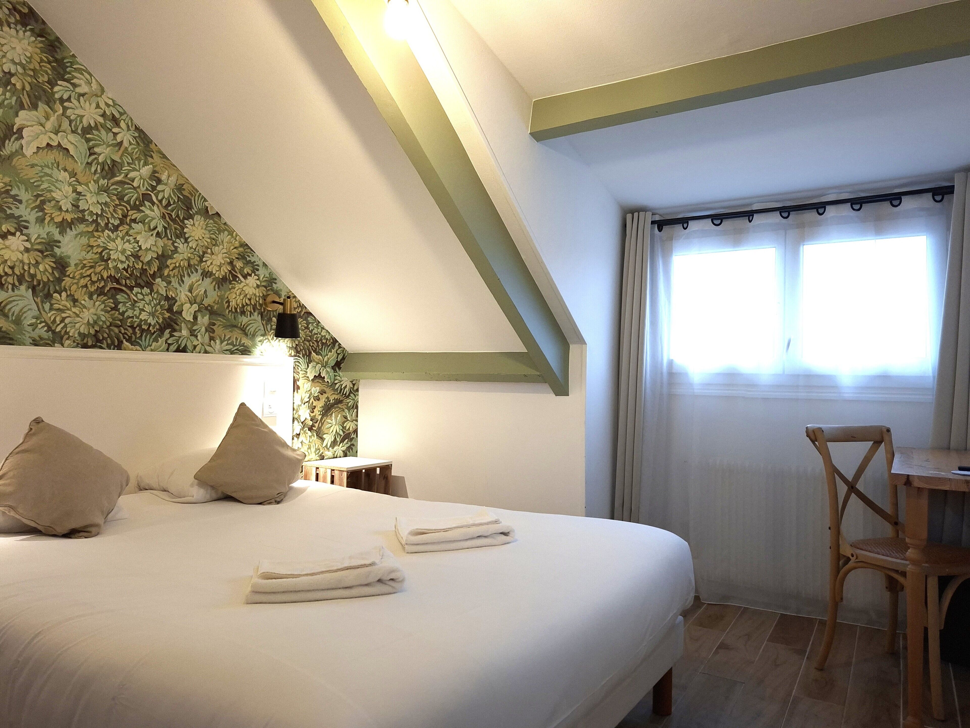 Une chambre à coucher avec un lit, des oreillers, une chaise, une table, une lampe et une fenêtre avec des rideaux.