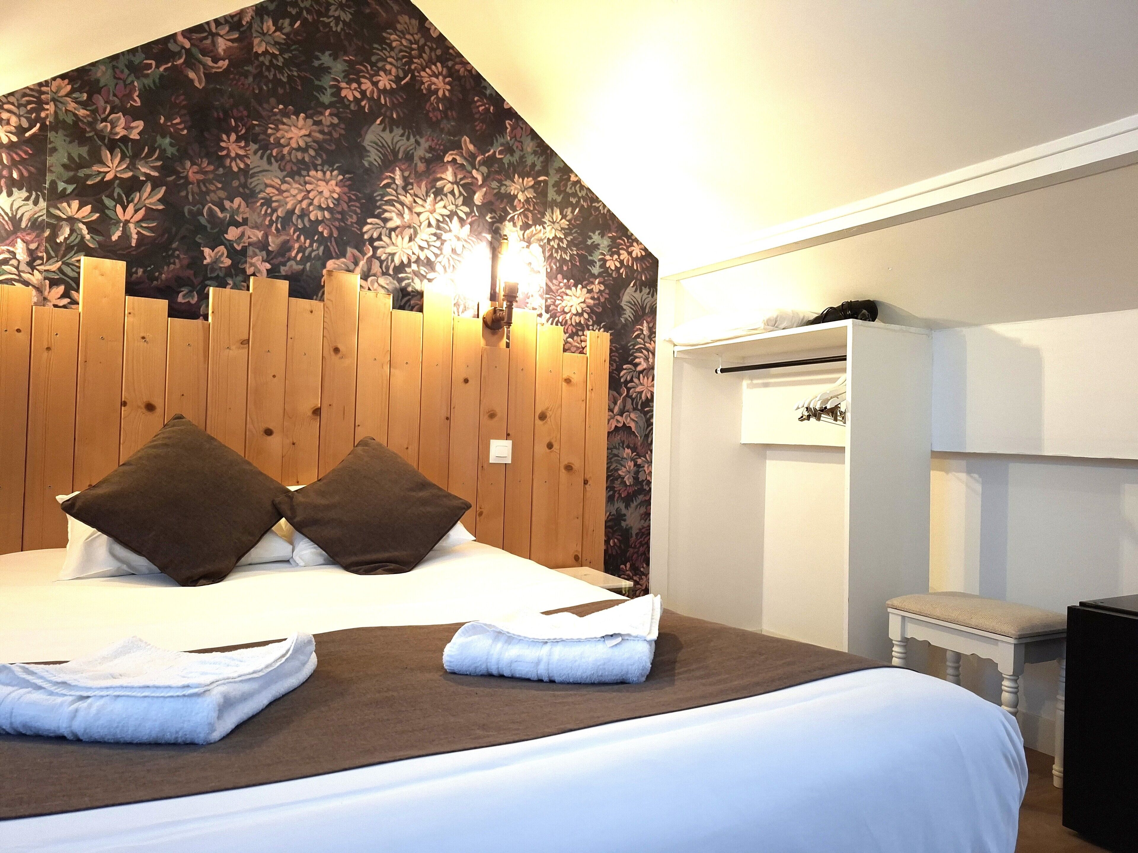 A hotel room with a wooden headboard, a bed with white linens, two brown pillows, and a small bedside table with a lamp.