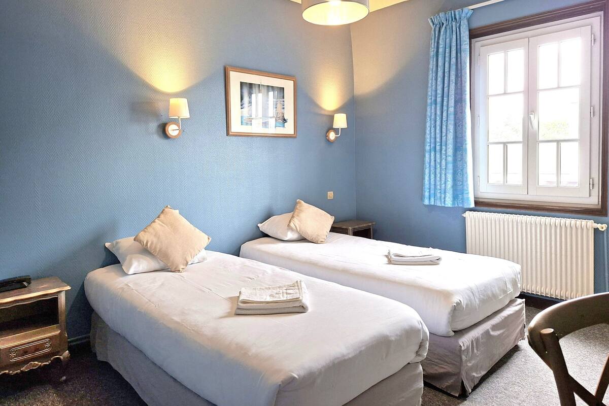 Une chambre avec deux lits, un tableau encadré au mur, une lampe et une fenêtre avec des rideaux.