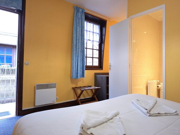Una habitación de hotel con cama, ventana con cortinas, radiador y una mesita.