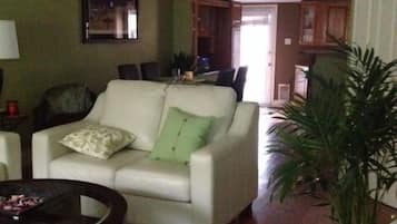 Un salon avec un canapé blanc, une table basse et une plante en pot. On aperçoit à l’arrière-plan un coin repas avec une table et des chaises.