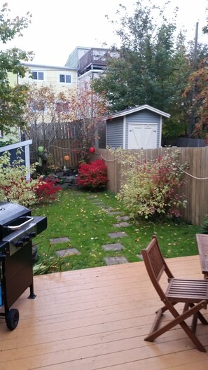 A backyard with a wooden deck, a barbecue grill, a shed, and various shrubs and trees. - Rice House Vacation Rental (St. John's)