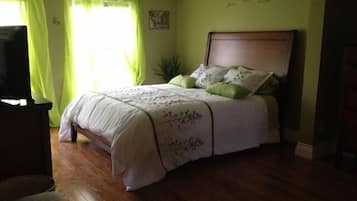 Une chambre avec un plancher en bois, un lit avec une literie blanche et des oreillers verts, un rideau vert et un mur orné d’un petit tableau.