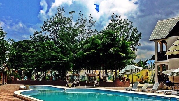 A rectangular swimming pool with a tiled deck, surrounded by lounge chairs and umbrellas, with trees and a building in the background. - Hotel Taciana (Léogâne)