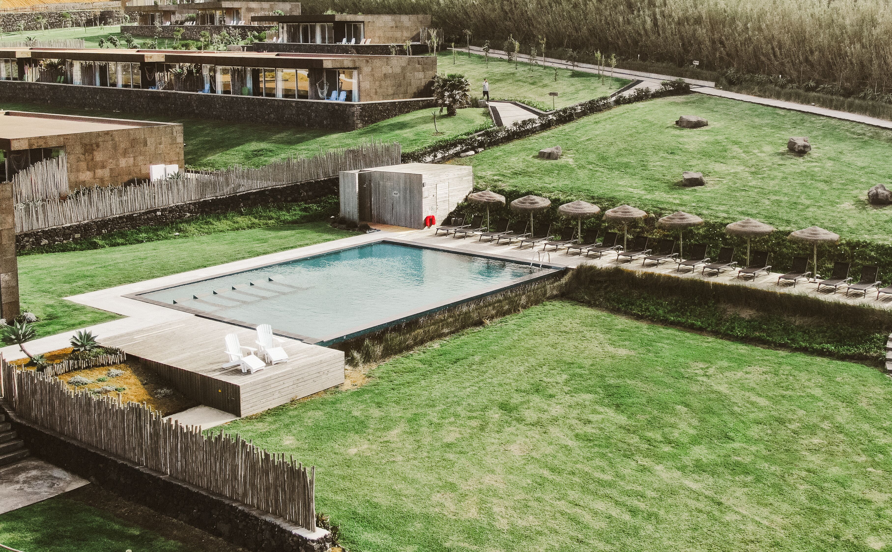 A swimming pool surrounded by lounge chairs and umbrellas, with a grassy area and a building in the background.