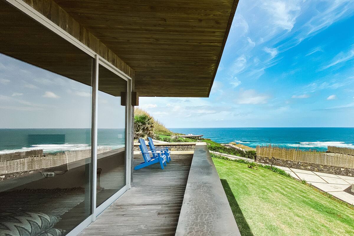 Een modern huis met een glazen deur die uitkomt op een balkon met uitzicht op de oceaan.