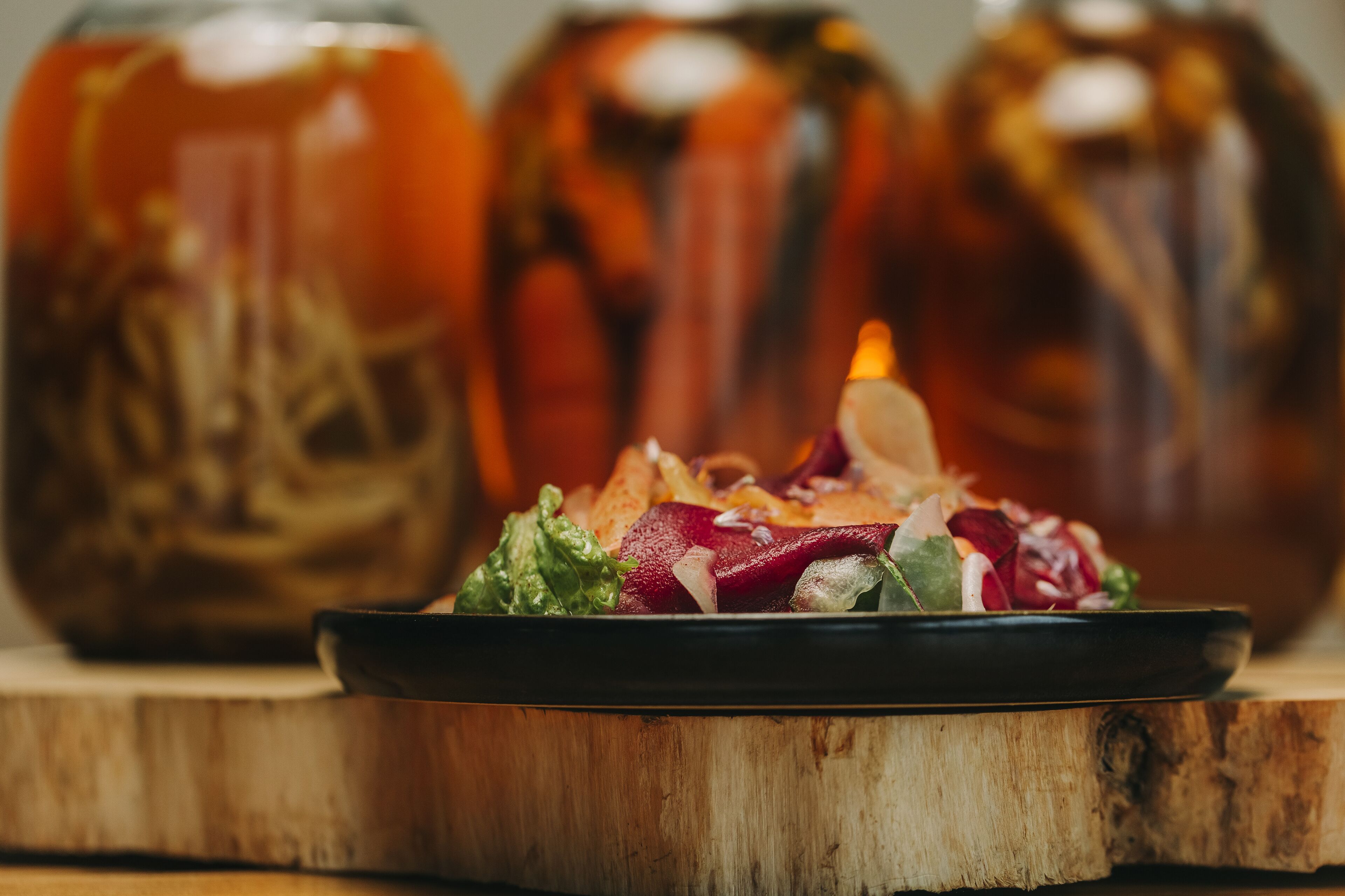 A wooden table with a black plate containing a salad. In the background, there are three glass jars with a golden liquid.